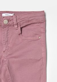 Jeans color malva chiaro con una texture liscia, dotati di una tasca frontale, chiusura con bottone metallico e dettagli cuciti sulle cuciture.