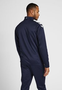Marineblauwe sportjacket met hoge kraag, lange mouwen en witte accenten op de schouders. Gemaakt van glad, lichtgewicht materiaal.