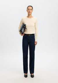 Črtasta smetanasta in bež majica s dolgimi rokavi, hlače z visokim pasom temno modre barve, črn pas, drži črno strukturirano torbico, obuta v ravne črne čevlje.