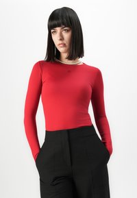 Vrouw met steil zwart haar, gekleed in een rode lange mouw top, zwarte broek en een parelketting, staande met handen in de zakken.