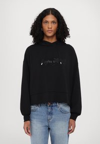 Black hooded sweatshirt with a cropped design, oversized sleeves, and a textured logo on the front. Worn with light blue jeans.