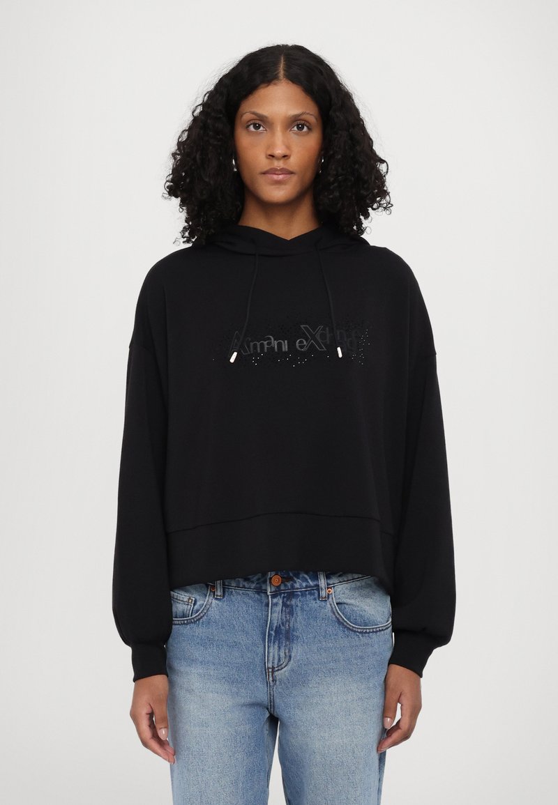 Black hooded sweatshirt with a cropped design, oversized sleeves, and a textured logo on the front. Worn with light blue jeans.