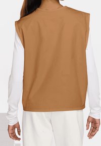 Tan bezramenný top s kulatým výstřihem, vyrobený z hladké látky, vrstvený přes bílou košili s dlouhým rukávem, s uvolněným střihem a čistými okraji.