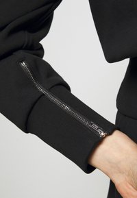 Černý rukáv mikiny s žebrovanou strukturou a stříbrným zipem podél manžety pro nastavitelný střih.