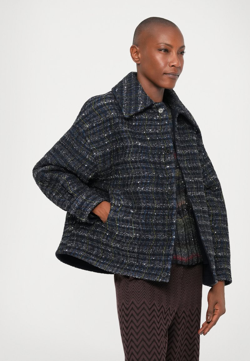 Marineblaues Tweed-Jacket mit großem Kragen, strukturiertem Muster, Knopfverschluss und Seitentaschen. Über gemusterten Hosen getragen.