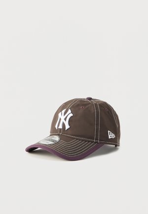 Tummanruskea New York Yankees -baseballlippalakki, jossa valkoinen logo, violetti lippareunus ja säädettävä hihna valkoisella taustalla.