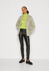 Ljusgrå blazer med en skräddarsydd passform, parat med en genomskinlig, ljusgrön polotröja och svarta smala byxor i konstläder. Svarta loafers fulländar looken.