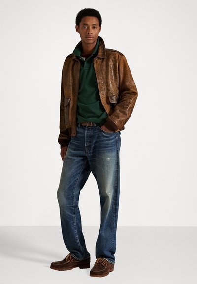 Bruine leren bomberjack, groene polo, versleten blauwe spijkerbroek, bruine veterschoenen met tan accenten, staand tegen een neutrale achtergrond.