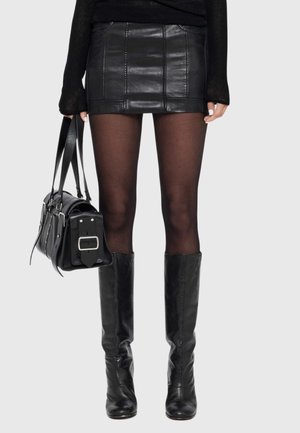 Femme portant une mini-jupe en cuir noir, des collants noirs transparents, des bottes noires montantes, tenant un sac à main en cuir noir avec une grande boucle.