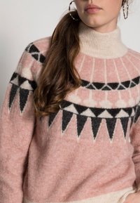 Pullover mit hohem Kragen, hergestellt aus flauschigem pinkem Material mit schwarz-weißen geometrischen Mustern. Das Design umfasst dreieckige Akzente.