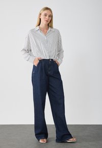 Hvit skjorte med svarte vertikale striper, kombinert med mørkeblå vide jeans og hvite sandaler, med hendene i lommmene.