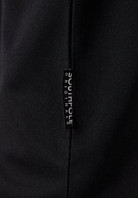 Schwarzer Stoff mit einer strukturierten Oberfläche, der ein kleines weißes Etikett mit der Aufschrift "SOU IT POLE" und "ORIGINALS" in einer kontrastierenden Schriftart trägt.