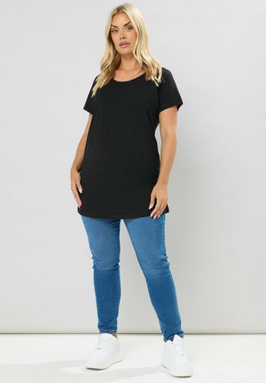 Mujer rubia de pie, vestida con una blusa negra holgada de manga corta, vaqueros ajustados azules y zapatillas blancas, sobre un fondo claro y liso.