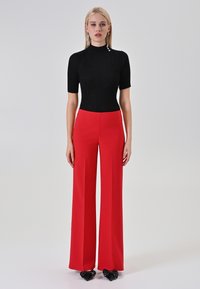 Top nero a maniche corte a coste abbinato a pantaloni rossi a vita alta con gamba larga. La modella indossa tacchi neri con fiocco. Sfondo semplice.