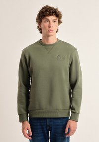 Sudadera verde olivo hecha de tela suave, con cuello redondo, puños acanalados y un logotipo circular en el pecho. Textura suave en general.