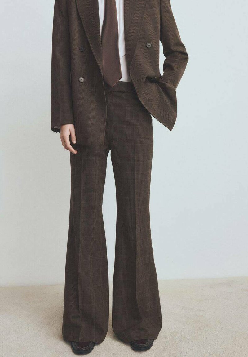 Osoba w brązowym w kratę garniturze z szerokimi spodniami, dopasowanym dwurzędowym żakiecie, białej koszuli i brązowym krawacie na jednolitym tle.