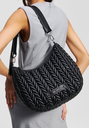 Vrouw in grijze mouwloze jurk met een grote zwarte doorgestikte schoudertas met brede riem en zichtbaar merkembleem.