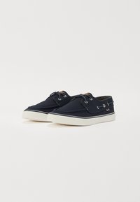 Zapatos de barco de lona azul marino con suelas de goma blanca, cordones y ojales de metal. Presentan un pequeño acento de bandera en el lado. Textura suave.