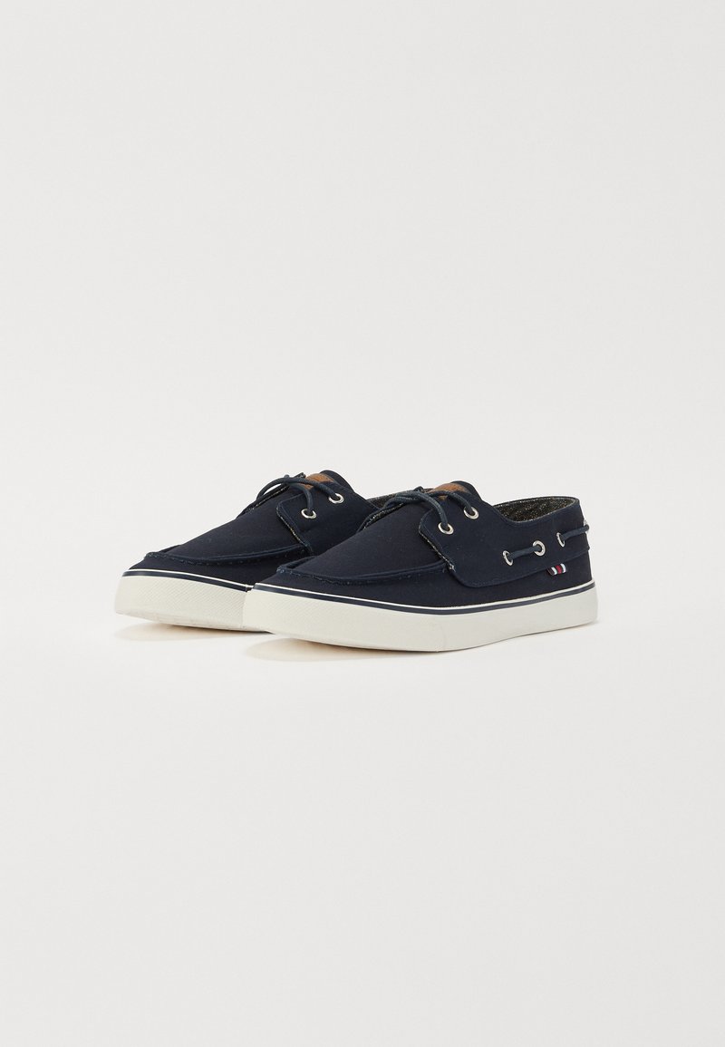 Zapatos de barco de lona azul marino con suelas de goma blanca, cordones y ojales de metal. Presentan un pequeño acento de bandera en el lado. Textura suave.