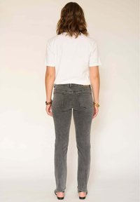 Femme aux cheveux bruns mi-longs portant une chemise blanche à manches courtes, un jean gris et des sandales noires, debout de dos sur un fond uni.