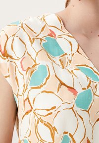 Blouse à manches courtes présentant un motif abstrait multicolore avec des formes turquoise, pêche et corail sur un fond crème.