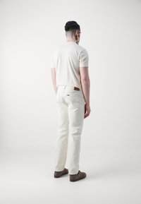 Cămașă scurtă de culoare bej deschis, asociată cu pantaloni albi. Pantalonii au buzunare pe spate și o etichetă din piele maro pe talie. Pantofi maro.