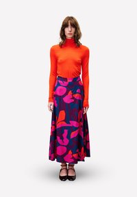 Langärmliges oranges Oberteil kombiniert mit einem schwingenden dunkelblauen Rock, der mit einem auffälligen rosa Blumenmuster verziert ist; Modell trägt schwarze Schuhe.