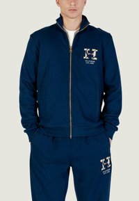 Giacca blu navy con zip, collo alto, cerniera dorata e logo decorativo sul petto. Abbinata a pantaloni coordinati. Materiale in misto cotone.