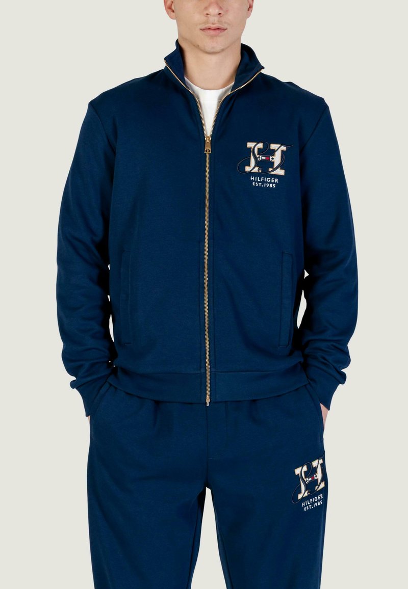 Giacca blu navy con zip, collo alto, cerniera dorata e logo decorativo sul petto. Abbinata a pantaloni coordinati. Materiale in misto cotone.