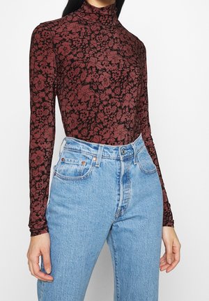 Vrouw draagt een zwart-wit botanische top met lange mouwen en hoge hals, gecombineerd met lichtblauwe high-waisted jeans tegen een effen achtergrond.