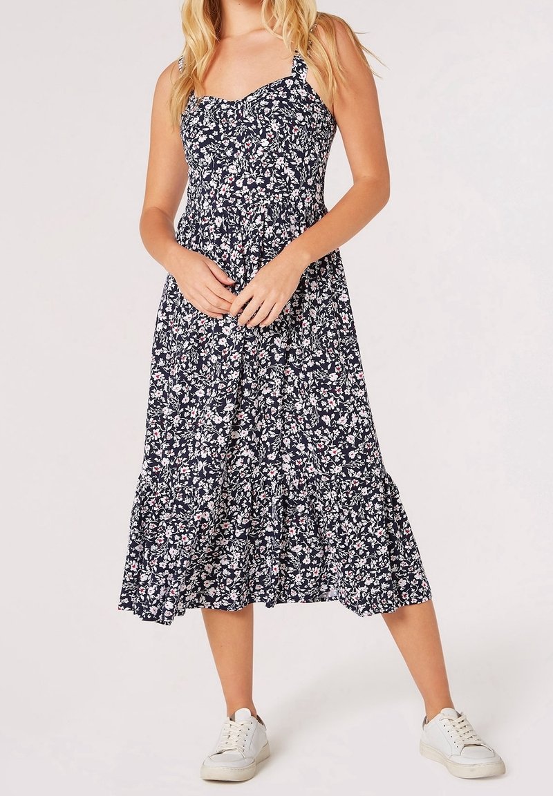 Femme portant une robe midi fleurie bleu marine à fines bretelles et des baskets blanches, debout, les mains jointes devant elle.