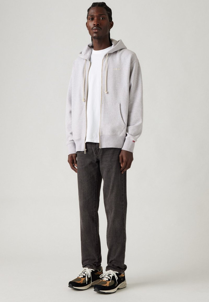 Sudadera gris con cremallera y cordones sobre una camiseta blanca, combinada con pantalones gris oscuro y zapatillas negras con acentos de camuflaje.