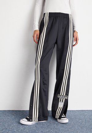 Osoba nosząca czarne spodnie dresowe Adidas z białymi paskami i zatrzaskami po bokach, w połączeniu z czarno-białymi sneakersami.