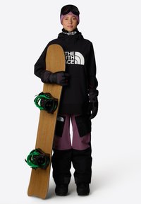 Snowboard med en träfot, gröna bindningar och svart hårdvara. Modellen bär en svart sweatshirt, lila byxor och svarta handskar, samt goggles.