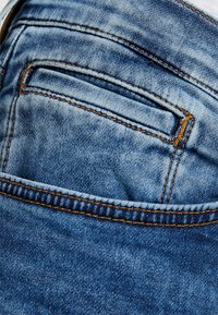 Jasnoniebieskie dżinsy z tylną kieszenią z kontrastowym pomarańczowym przeszyciem i wyblakłą teksturą, podkreślające swobodny styl.