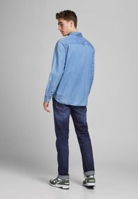 Giovane uomo in piedi con le spalle girate, indossando una camicia di jeans blu chiaro, jeans blu scuro e sneaker verdi e bianche su uno sfondo grigio.