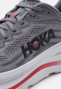 Γκρι αθλητικό παπούτσι HOKA με υφαντό ύφασμα στο πάνω μέρος, γκρι κορδόνια, μαύρο λογότυπο με κόκκινη λεπτομέρεια και λευκή σόλα με κόκκινη ρίγα.