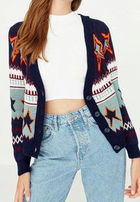 Strickjacke in Marineblau mit bunten Stern- und geometrischen Mustern. Mit Knopfverschluss und gerippten Bündchen. Über einem weißen Oberteil getragen.