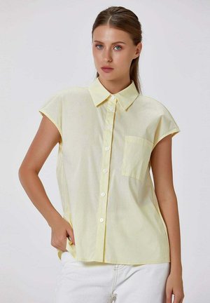 Femme portant une chemise jaune clair à manches courtes avec une poche poitrine, posant avec une main dans la poche d'un pantalon blanc.