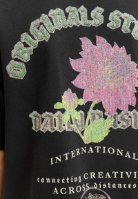 Melna T-krekli, kas satur daudzkrāsainus ziedu grafiskos attēlus un teksturētas burtu atveidojumus dažādās zaļās un rozā nokrāsās.