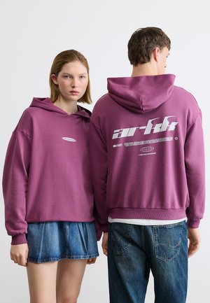 Two young people, one facing front in a purple hoodie and denim skirt, the other facing back in a matching hoodie and blue jeans.