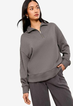 Šedá mikina s polo zipem, oversize střih a žebrované manžety. Materiál vypadá měkký a texturovaný, stylizováno s uvolněnými kalhotami.