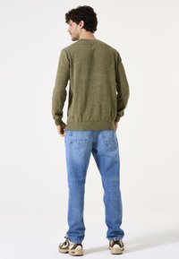 Grüner Strickpulli mit gerippten Bündchen und Saum, kombiniert mit hellblauen Jeans mit Gesäßtaschen und einer lockeren Passform. Legere sportliche Sneakers.