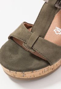 Sandale plateforme en suede vert olive avec un talon en liège texturé et un large design de brides croisées cousues. Semelle intérieure lisse de couleur naturelle.