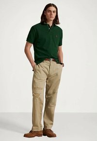 Πράσινο πουκάμισο πόλο με γιακά, μπεζ τζιν παντελόνι με φθορές και καφέ λόφρακες. Σημαντικά χαρακτηριστικά περιλαμβάνουν ένα μικρό λογότυπο στο στήθος.