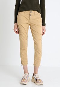 Osoba nosząca beżowe, krótkie spodnie cargo z zamkami, w połączeniu z wielokolorowymi tenisówkami i ciemnozielonym, prążkowanym, długim topem z długim rękawem.