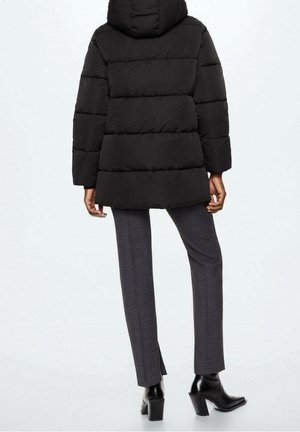Manteau matelassé noir à capuche, avec un motif matelassé, une longueur courte et des manches larges. Associé à un pantalon gris sur mesure et des bottes noires à talons.