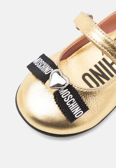 Goldene metallische Mary Jane Schuhe mit einem schwarzen elastischen Riemen, der weißen Markentext und einen silbernen herzförmigen Akzent an der Spitze besitzt.