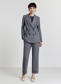 Abito grigio su misura doppiopetto con revers a punta, due tasche anteriori e pantaloni a gamba larga, abbinato a scarpe basse beige con punta a punta.