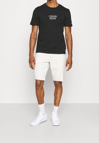 Camiseta de algodón negra con el logo "CALVIN KLEIN", combinada con pantalones cortos de mezclilla color blanco roto. El modelo lleva zapatillas blancas y calcetas tipo crew.